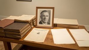 Table de musée présentant des dossiers d’archives anciennes, un portrait judiciaire masculin en noir et blanc, des feuilles de presse d’après-guerre et des documents d’enquête sous une lumière douce.