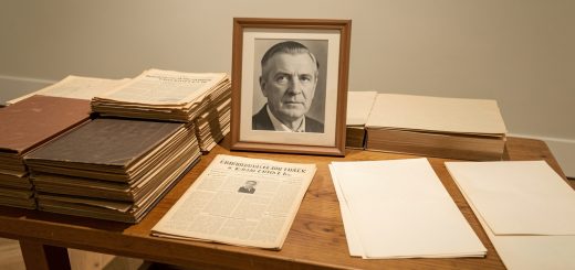Table de musée présentant des dossiers d’archives anciennes, un portrait judiciaire masculin en noir et blanc, des feuilles de presse d’après-guerre et des documents d’enquête sous une lumière douce.